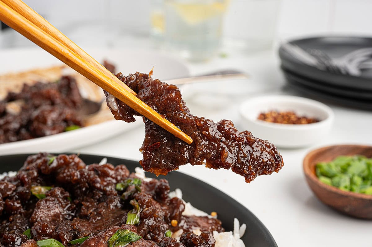 Chopsticks lifting a piece of glazed Mongolian beef above a bowl of beef and rice.