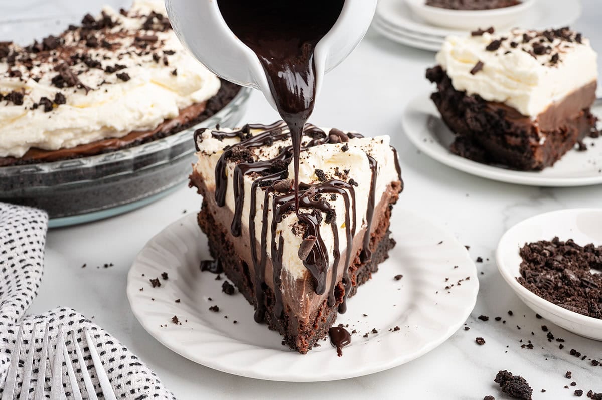 Chocolate sauce being drizzled over a slice of mud pie topped with whipped cream.