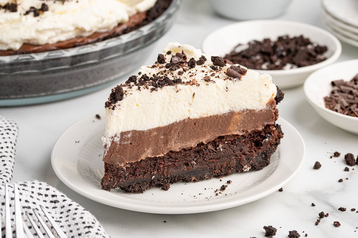 Slice of layered mud pie with chocolate crust, chocolate pudding, and whipped cream on a plate.
