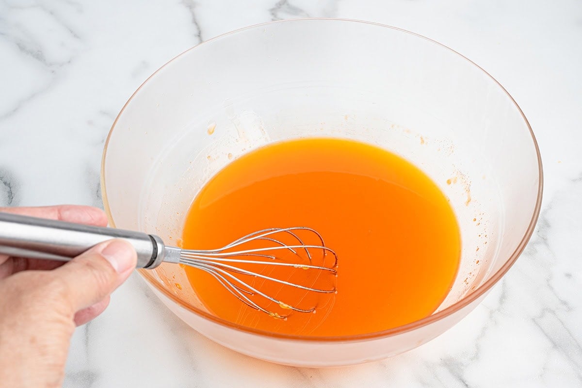 Hand whisking orange gelatin mixture in a clear mixing bowl on a marble countertop.