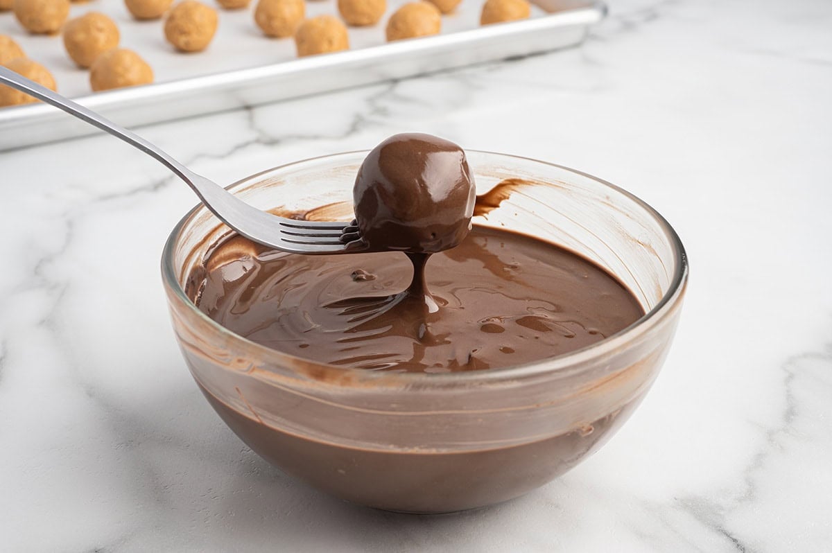 Peanut butter ball being dipped in melted chocolate with a fork over a glass bowl.