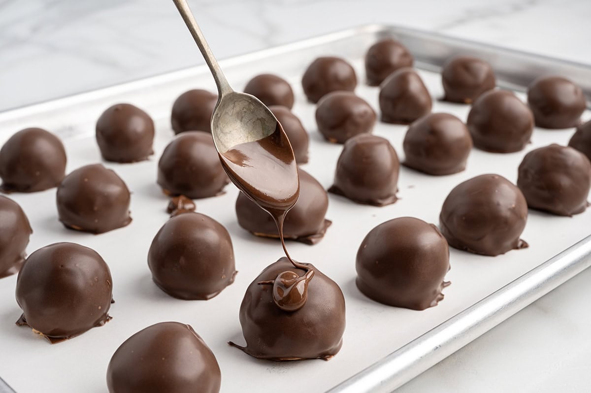 Drizzling melted chocolate over chocolate-dipped peanut butter balls on a tray.