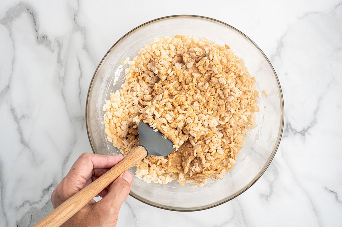 Hand folding rice cereal into peanut butter mixture with a spatula.