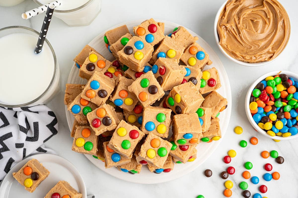 A plate piled high with peanut butter fudge squares, each topped with colorful M&M's candies, surrounded by bowls of peanut butter, extra M&M's, and glasses of milk.