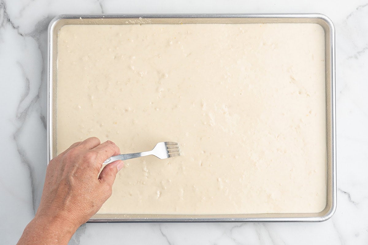 Hand docking rectangular pizza dough with a fork on a baking sheet.
