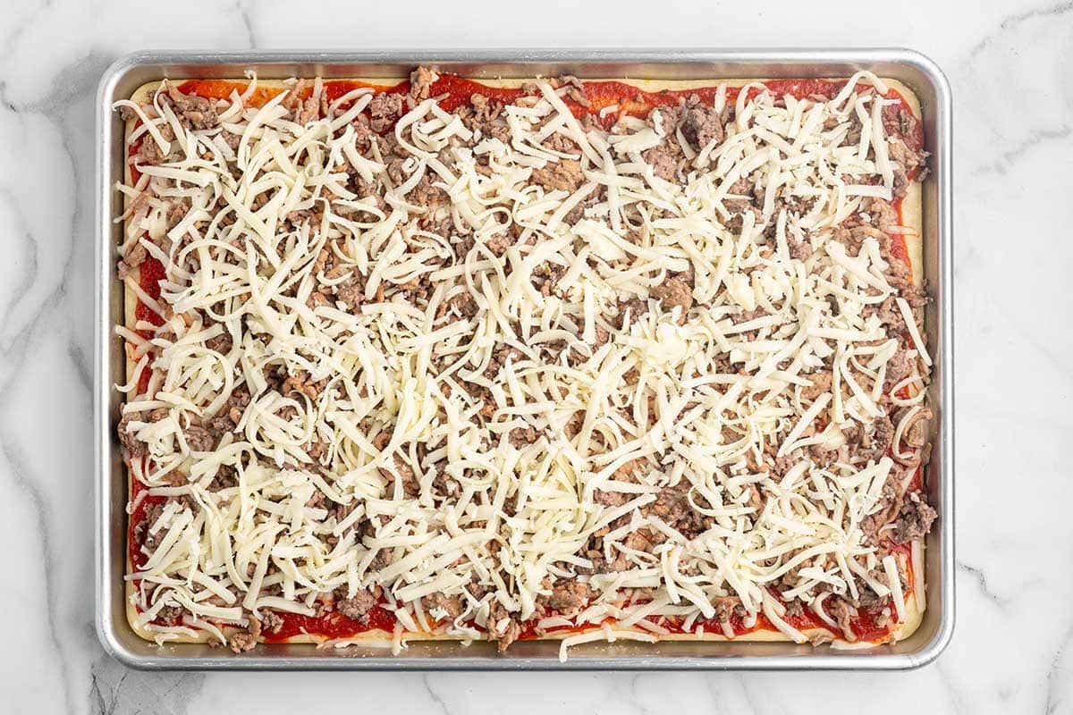Unbaked sheet pan pizza topped with sauce, sausage, and shredded mozzarella.