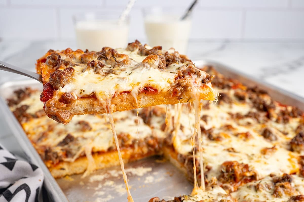 Slice of school pizza with melted cheese and sausage being lifted from the pan.