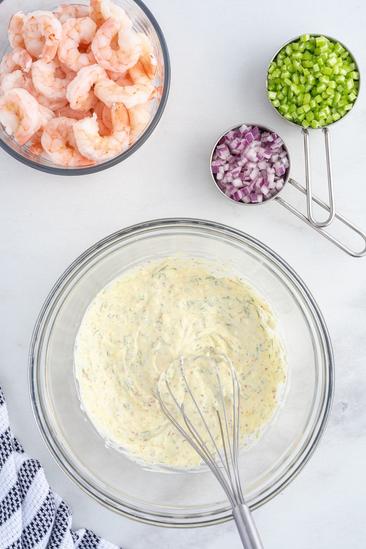 Shrimp Salad ingredients prepped overhead, creamy dressing in a glass bowl with a whisk, alongside cooked shrimp, diced celery, and red onion.
