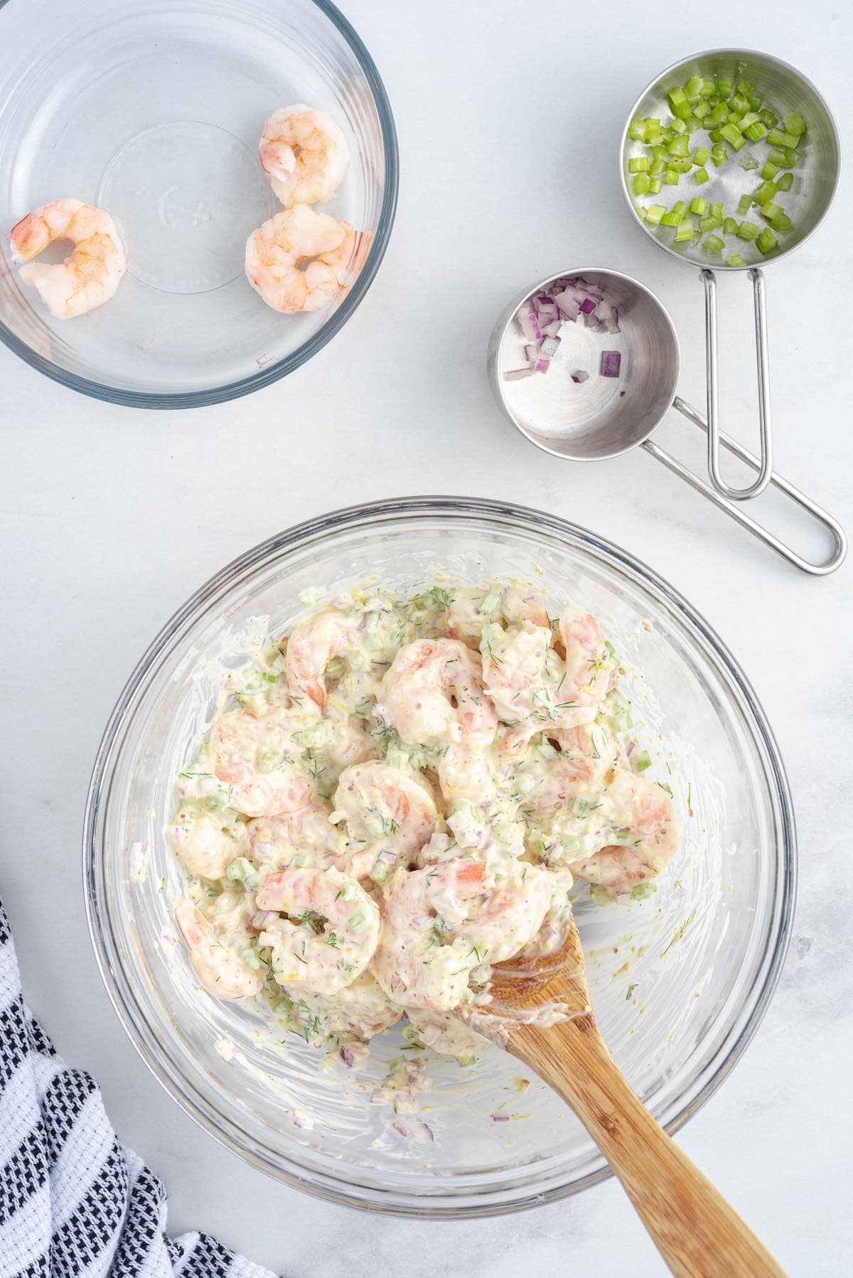 Shrimp Salad being mixed together in a glass bowl with a wooden spoon, with remaining shrimp, celery, and red onion alongside.