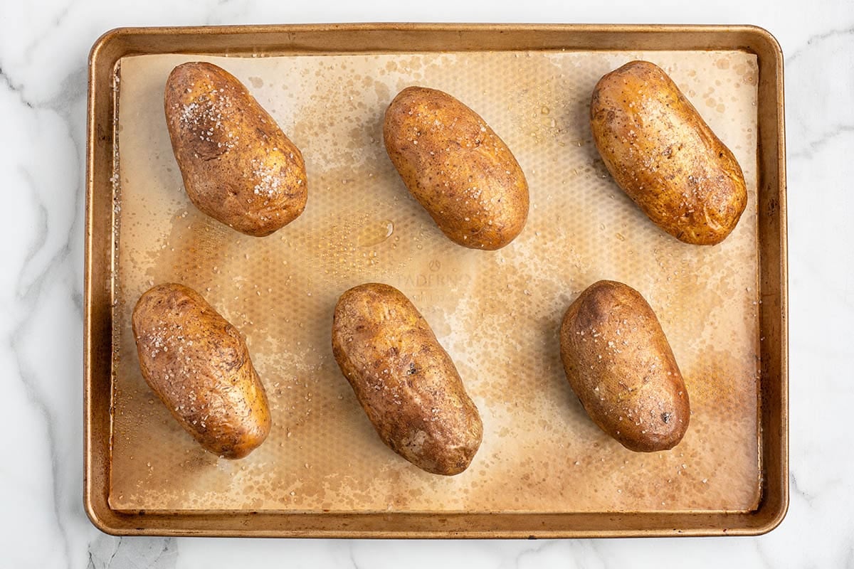 Six oil-rubbed and salted russet potatoes on a parchment-lined baking sheet ready to bake.