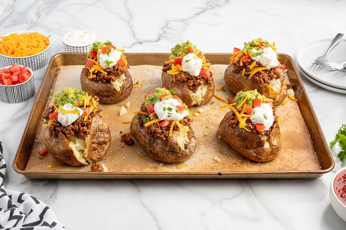 Six fully loaded Taco Potatoes on a baking sheet with seasoned ground beef, shredded cheddar, guacamole, sour cream, diced tomatoes, and cilantro.