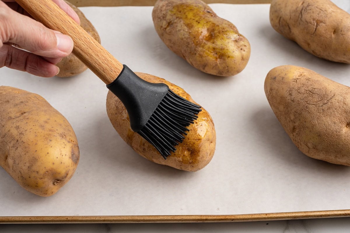 Brushing olive oil onto russet potatoes on a parchment-lined baking sheet before baking.