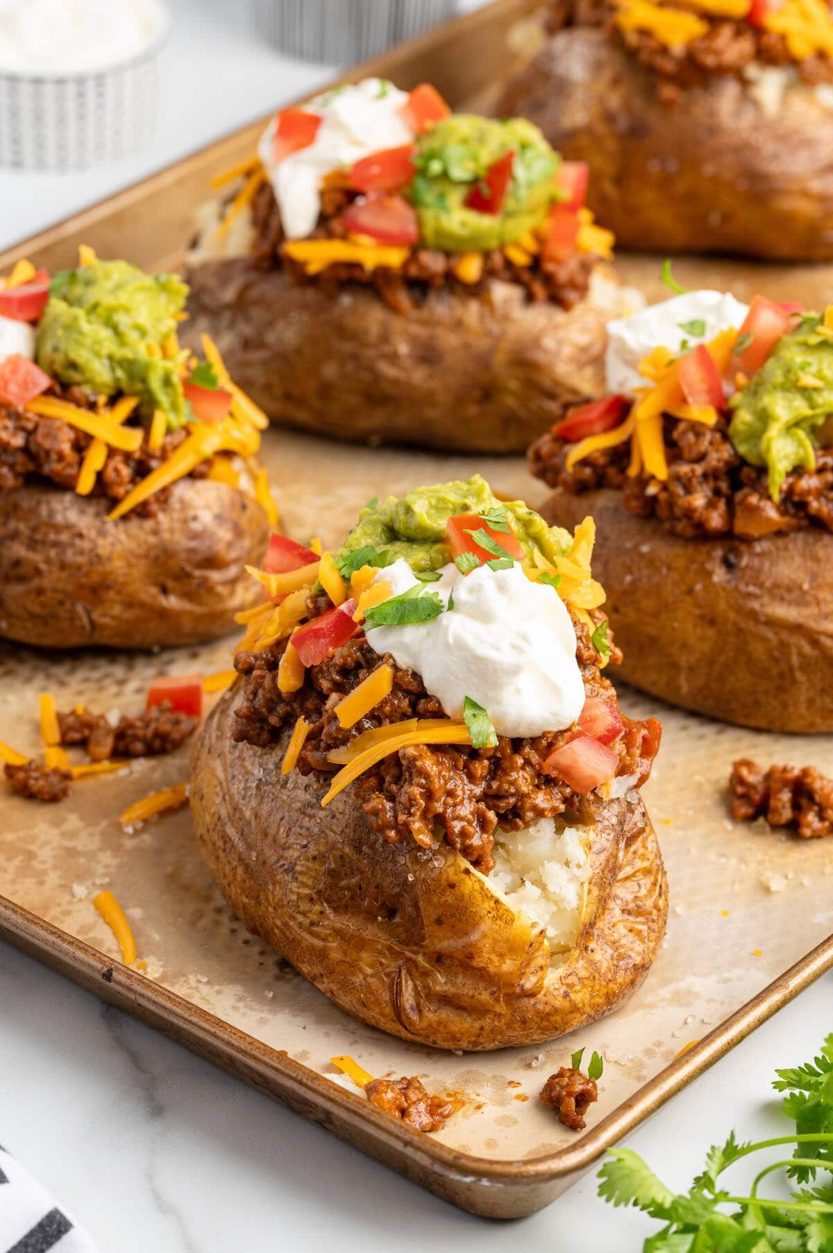 Four Taco Potatoes on a baking sheet topped with seasoned ground beef, shredded cheddar, guacamole, sour cream, diced tomatoes, and cilantro.