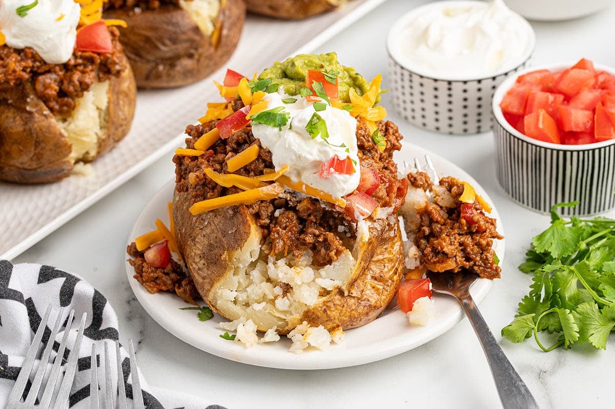 A Taco Potato being eaten with a fork on a white plate, loaded with seasoned ground beef, shredded cheddar, guacamole, sour cream, diced tomatoes, and cilantro.