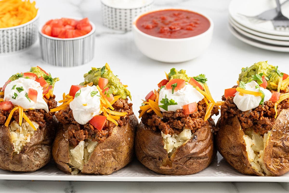 Four Taco Potatoes lined up on a white platter loaded with seasoned ground beef, shredded cheddar, guacamole, sour cream, diced tomatoes, and cilantro, with bowls of toppings and salsa in the background.