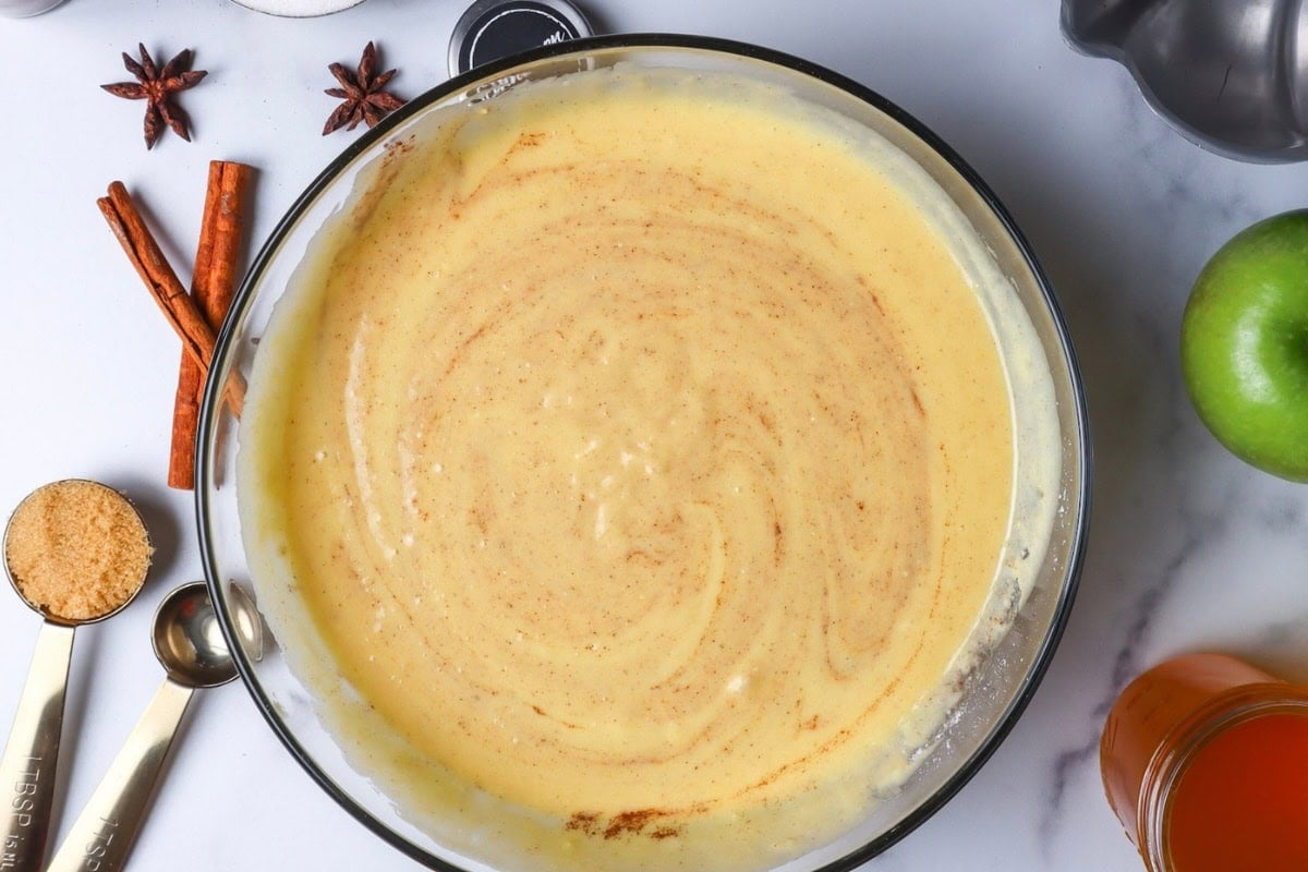 Cake batter with cinnamon swirls in a glass bowl, surrounded by cinnamon sticks, star anise, brown sugar, a green apple, and apple cider.