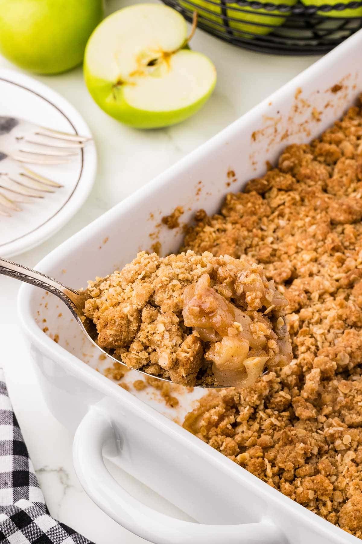Spoon lifting a serving of apple crisp with oat crumble topping from the baking dish.