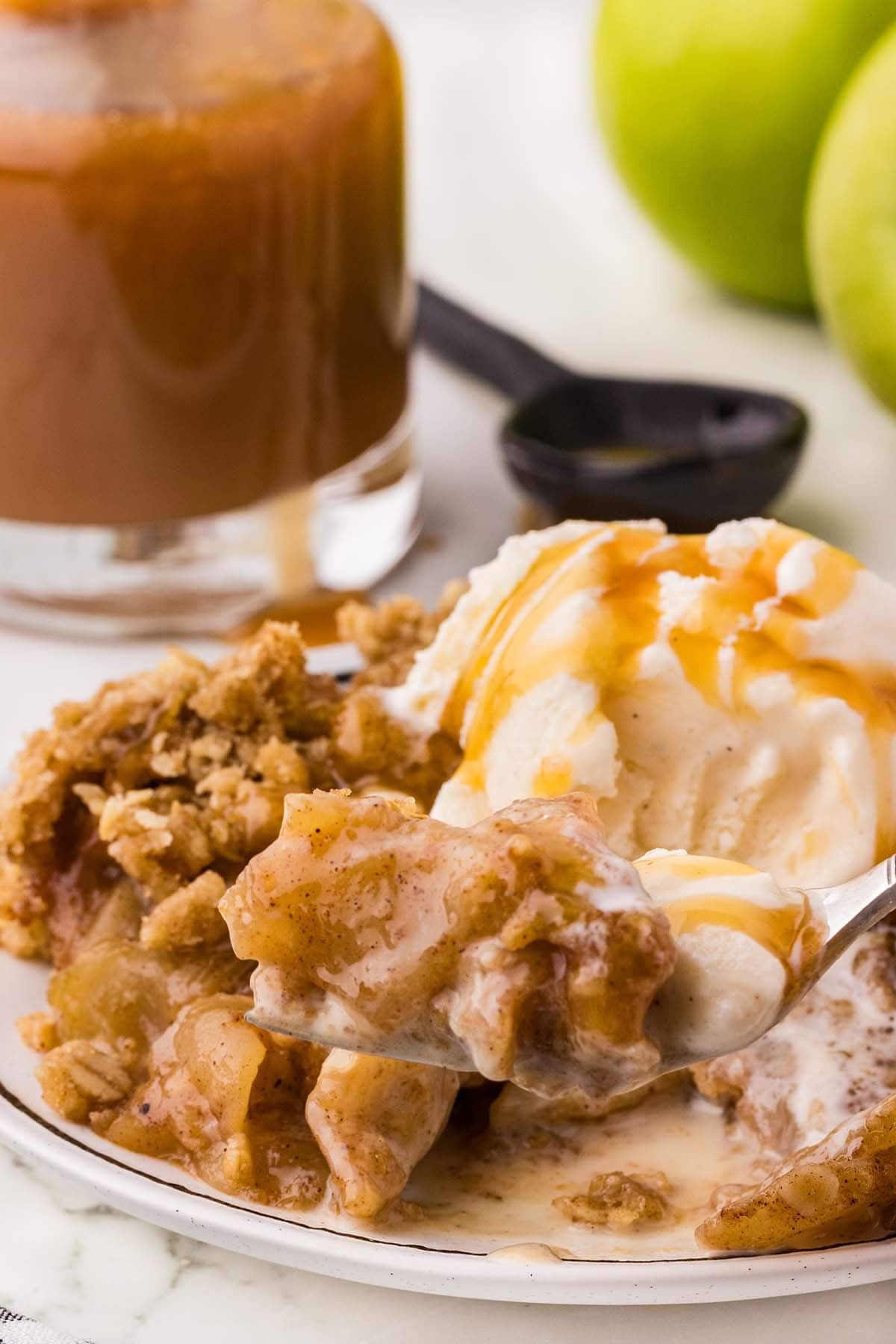 Forkful of apple crisp with vanilla ice cream and caramel drizzle held above the plate.