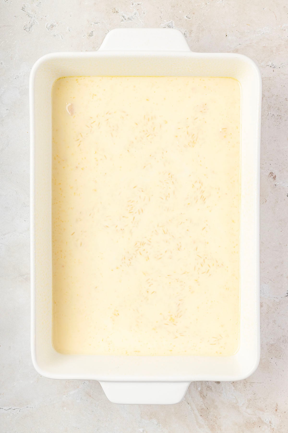 Overhead view of raw rice and creamy liquid in a white baking dish, ready to bake.