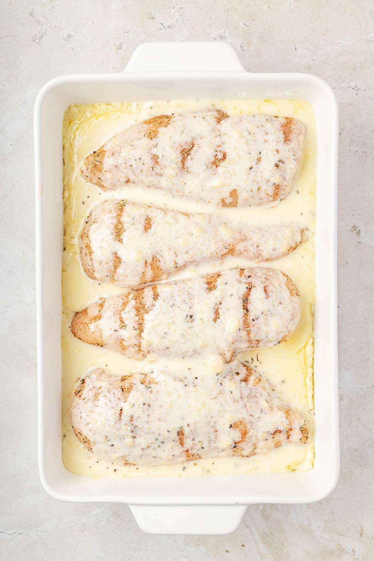 Overhead view of seasoned raw chicken breasts arranged over creamy rice mixture in a baking dish.