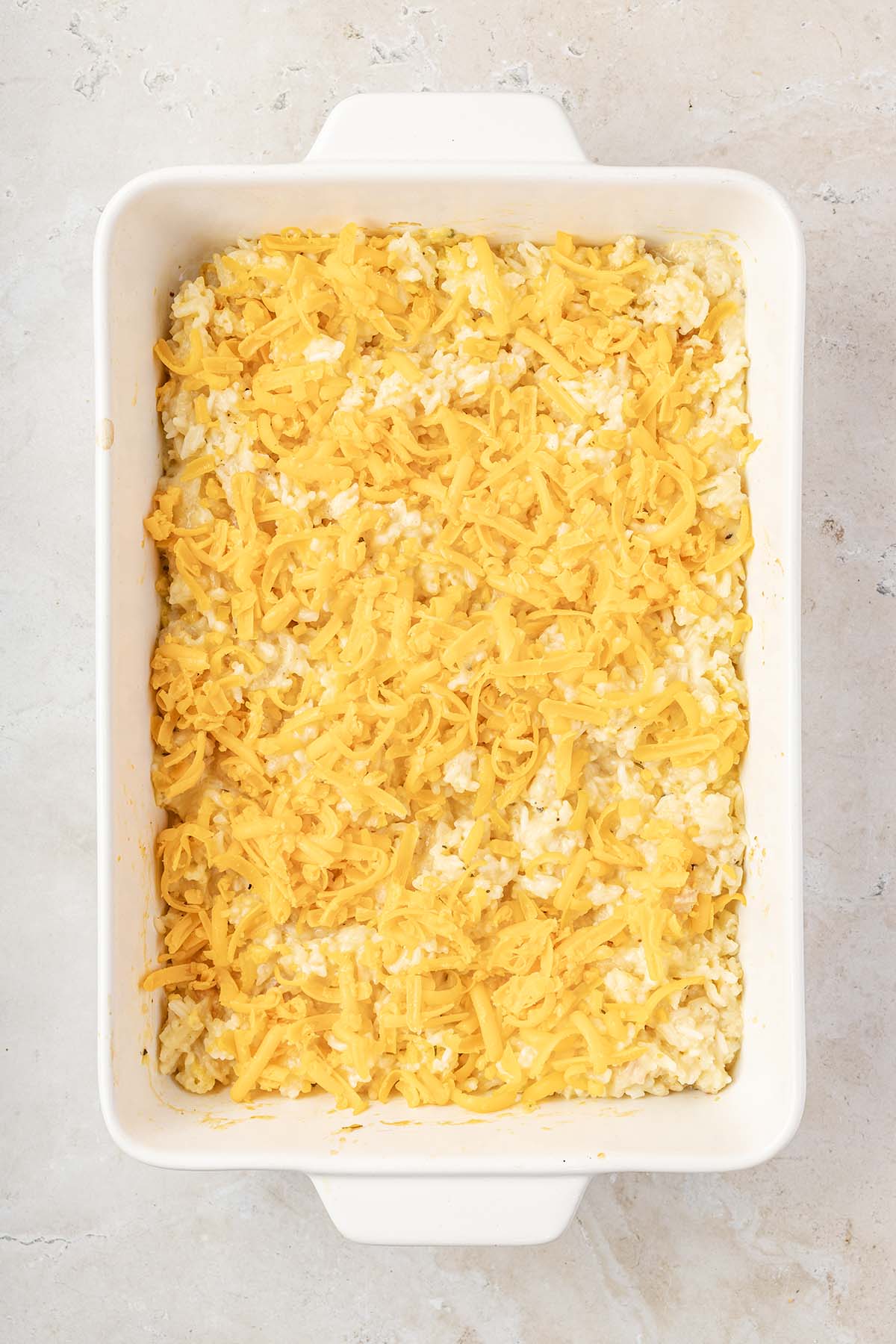 Overhead view of shredded cheese covering a creamy rice casserole in a white baking dish.