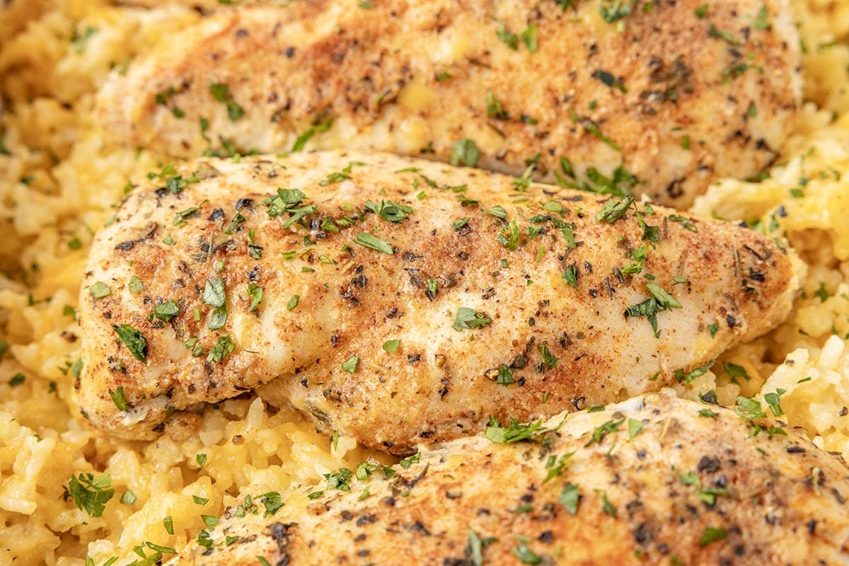 Close-up of seasoned baked chicken breasts resting on golden yellow rice, garnished with fresh herbs.