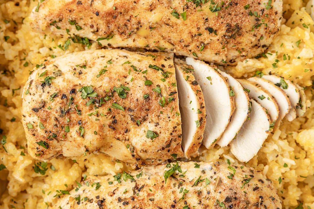 Close-up of sliced seasoned baked chicken breasts served over yellow rice, garnished with fresh herbs.