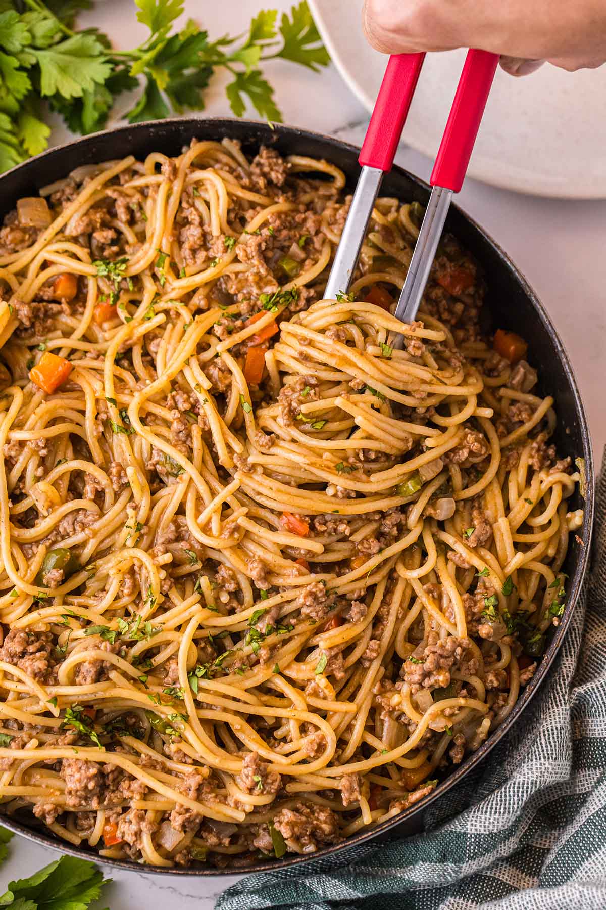Dirty Spaghetti with ground beef and vegetables being lifted with red pasta tongs.