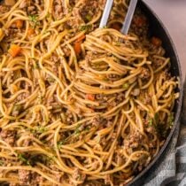 Close-up of tongs lifting dirty spaghetti with ground meat and vegetables from a skillet.
