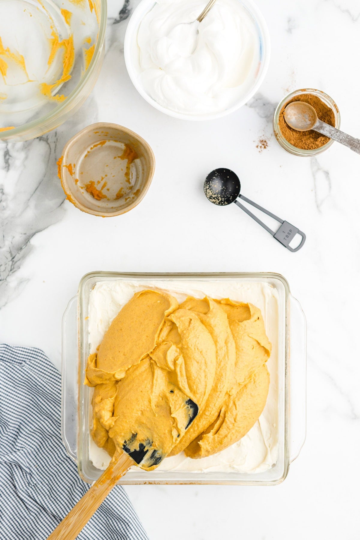 Spreading a pumpkin layer over the cream cheese layer in a baking dish, surrounded by whipped topping and cinnamon.
