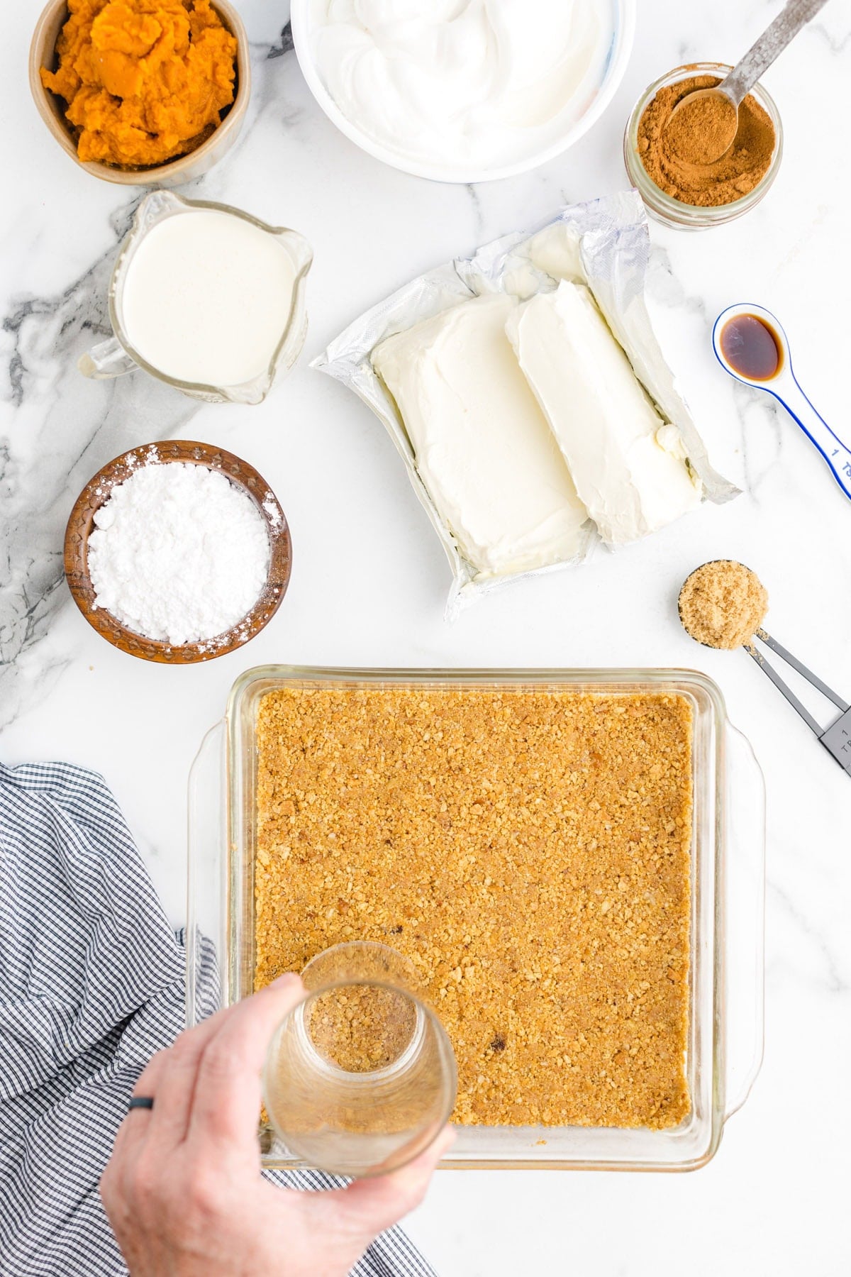 Pressing a graham cracker crust into a baking dish, surrounded by pumpkin puree, cream cheese, whipped topping, and other ingredients.