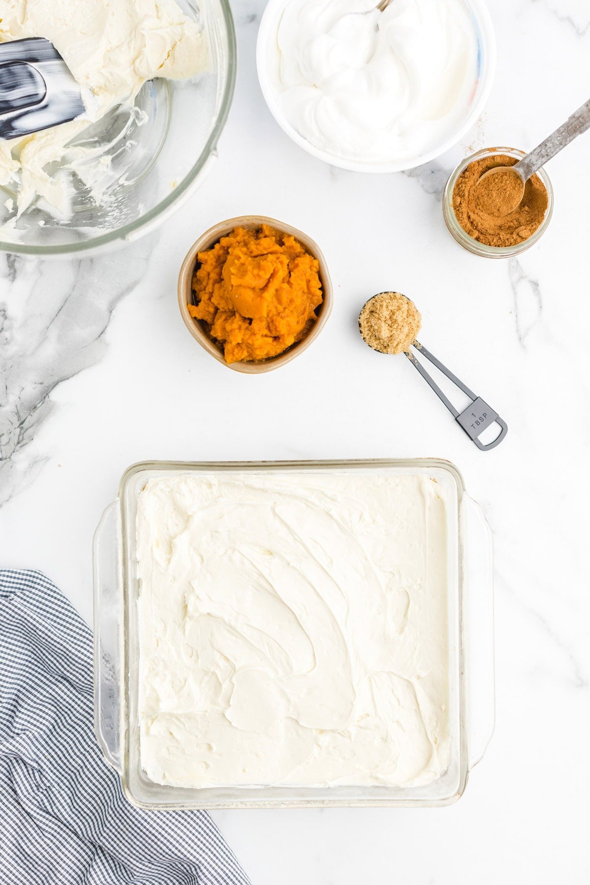 Cream cheese layer spread in a baking dish, surrounded by pumpkin puree, whipped topping, cinnamon, and brown sugar.