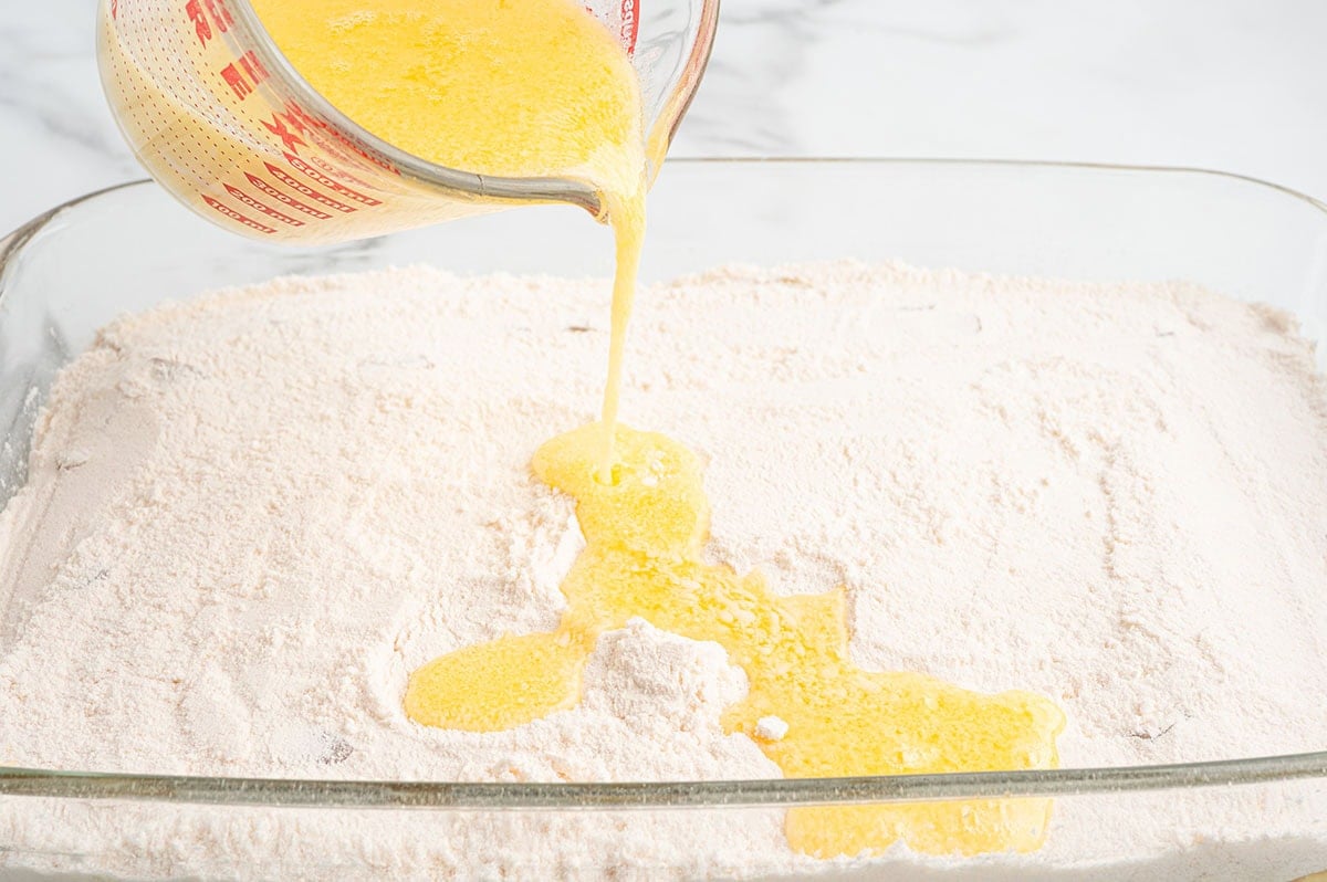 Pouring melted butter over dry cake mix layered on top of apples in a baking dish.