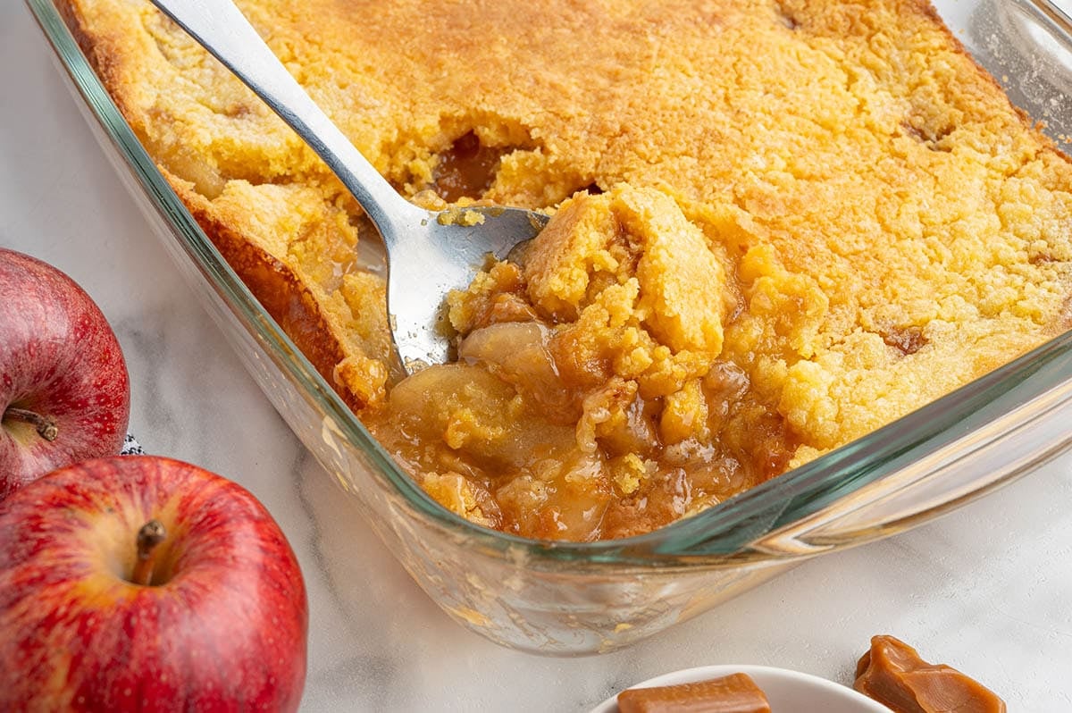 Spoon scooping caramel apple dump cake from a glass baking dish next to fresh apples.