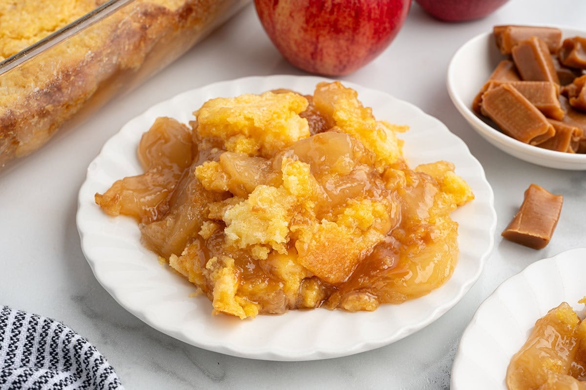 Plate of caramel apple dump cake with a golden cake topping and caramel apples nearby.