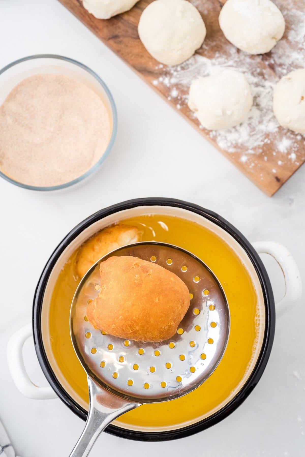 Fried apple pie bomb lifted from hot oil with a slotted spoon, dough balls and cinnamon sugar nearby