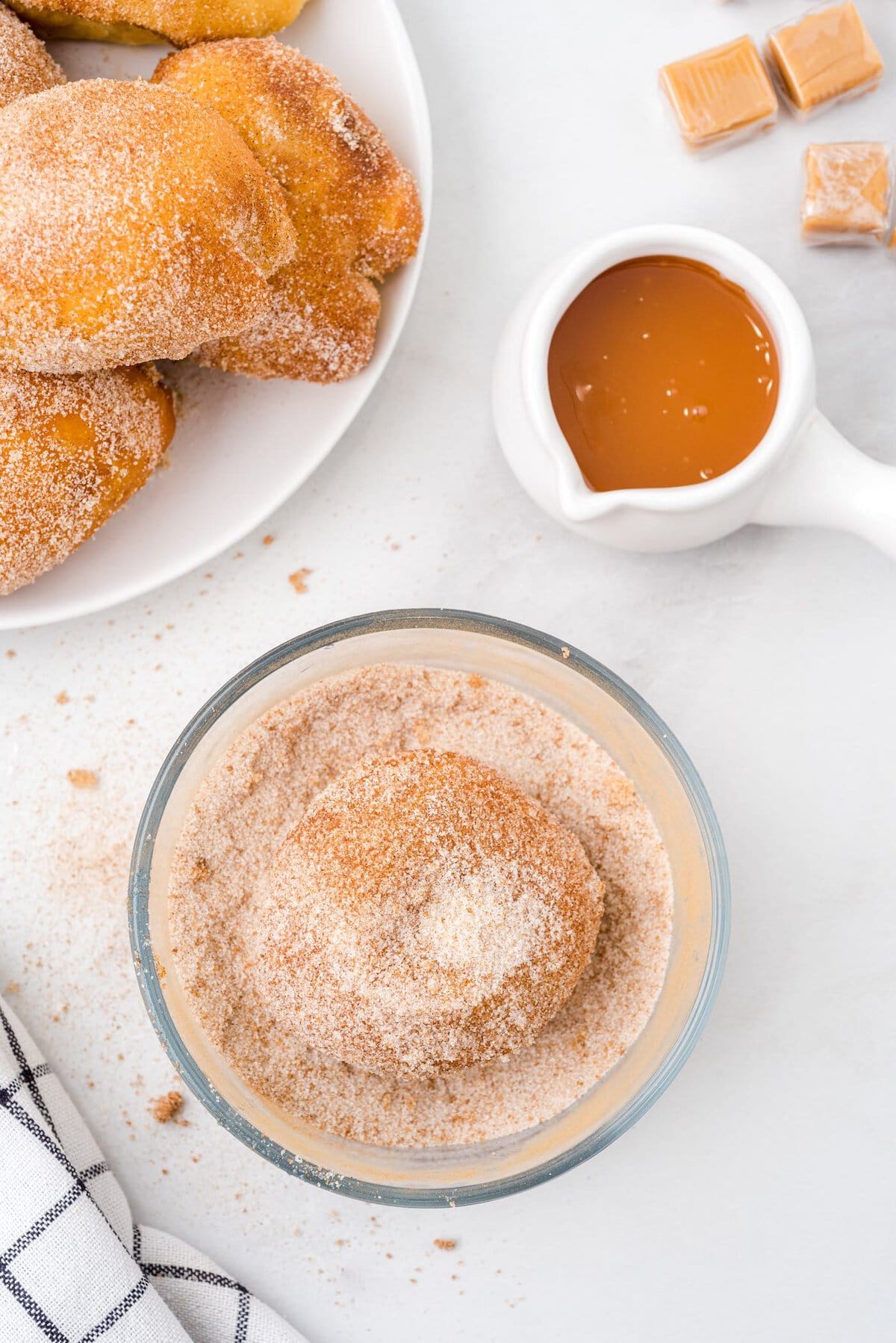 Caramel apple pie bomb being coated in cinnamon sugar next to a plate of finished bombs and caramel sauce
