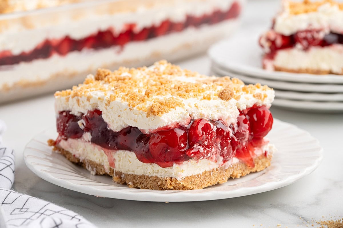 Layered cherry yum yum square on a plate, with the pan and another slice in the background.
