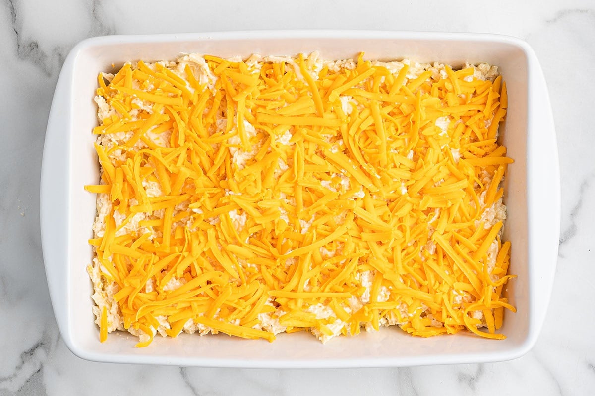 Hashbrown casserole topped with shredded cheddar cheese in a baking dish before baking.