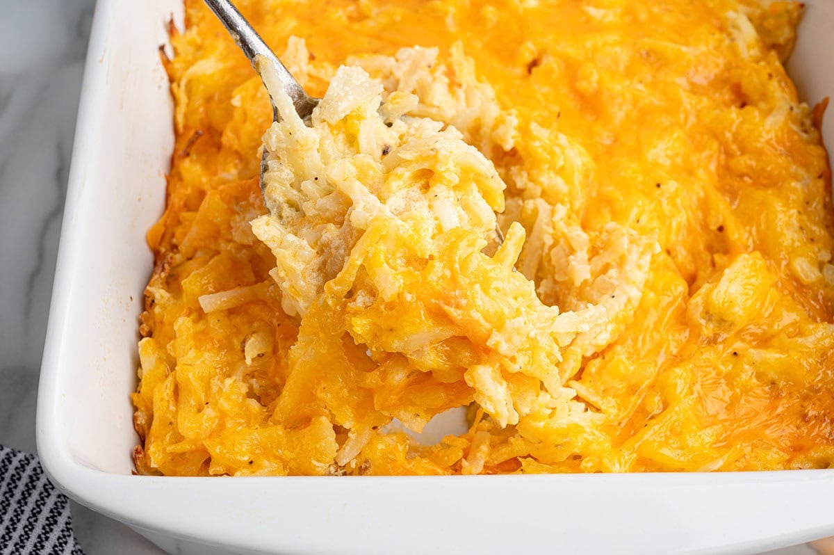 Spoon scooping cheesy hashbrown casserole from a white baking dish.