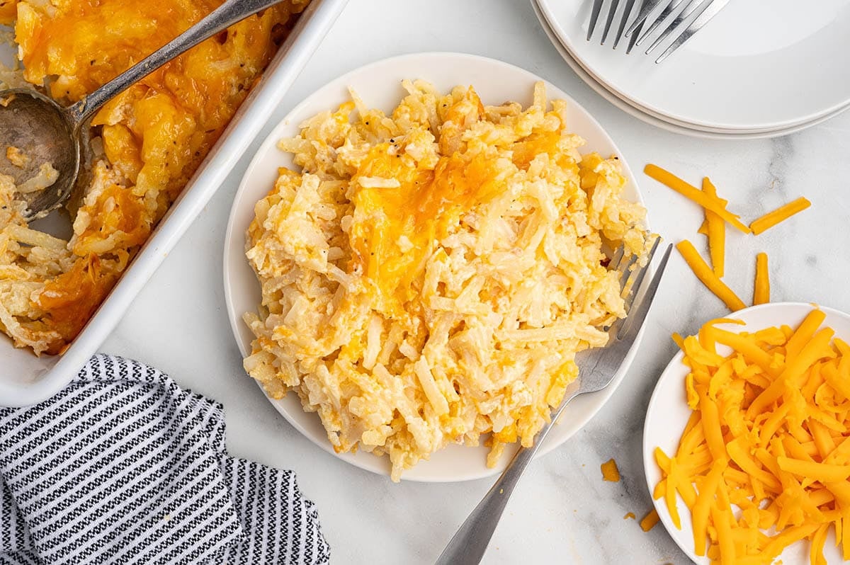 Plated cheesy hashbrown casserole with a fork, next to the baking dish and shredded cheese