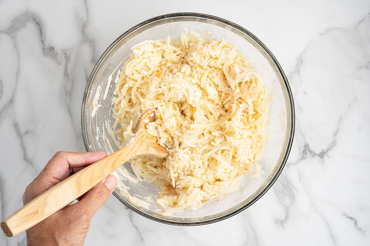Hand stirring shredded hashbrown casserole mixture in a glass bowl with a wooden spoon.