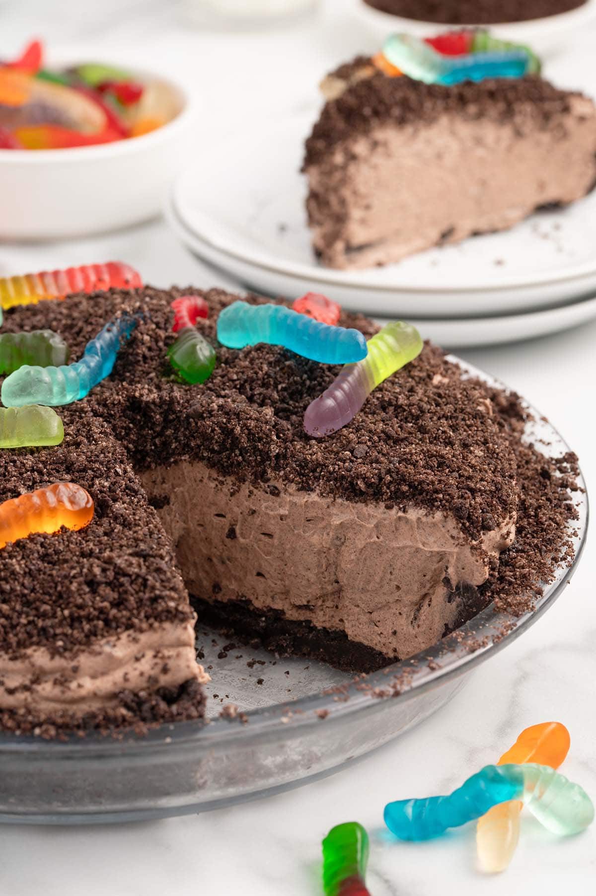 Dirt pie with a slice cut out, showing creamy filling and gummy worm topping, with a slice on a plate behind it.