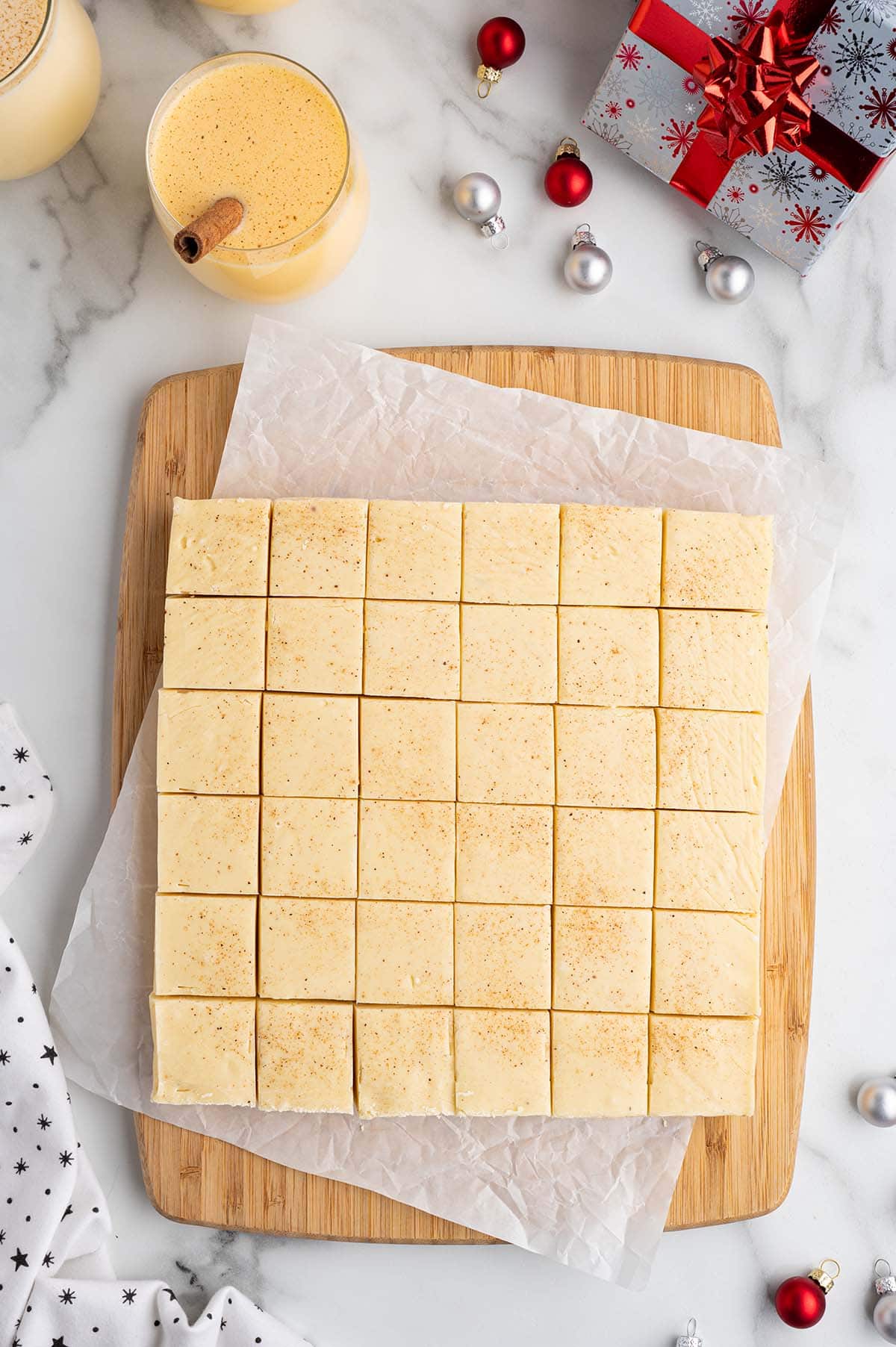 Eggnog fudge squares arranged on a cutting board with eggnog and Christmas decorations.