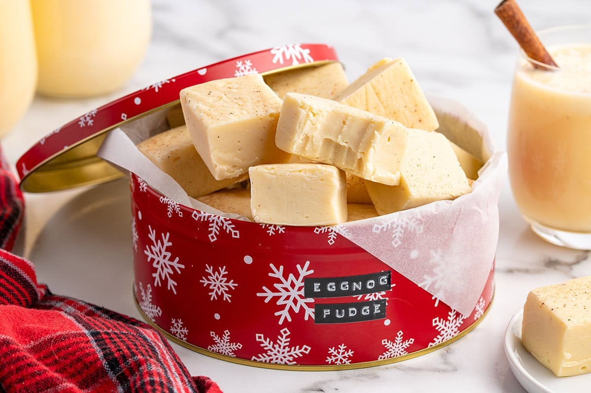 Eggnog fudge squares served in a festive red Christmas tin with a glass of eggnog.