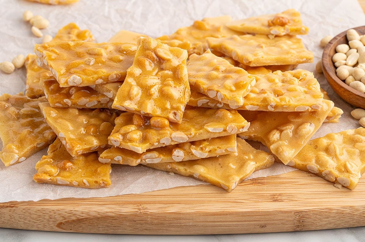 Broken pieces of peanut brittle stacked on a wooden cutting board.