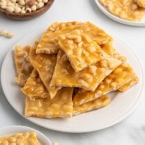 Pieces of peanut brittle piled on a white plate with a bowl of peanuts nearby.