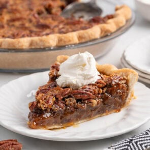Slice of pecan pie with whipped cream on a white plate, whole pie in the background.