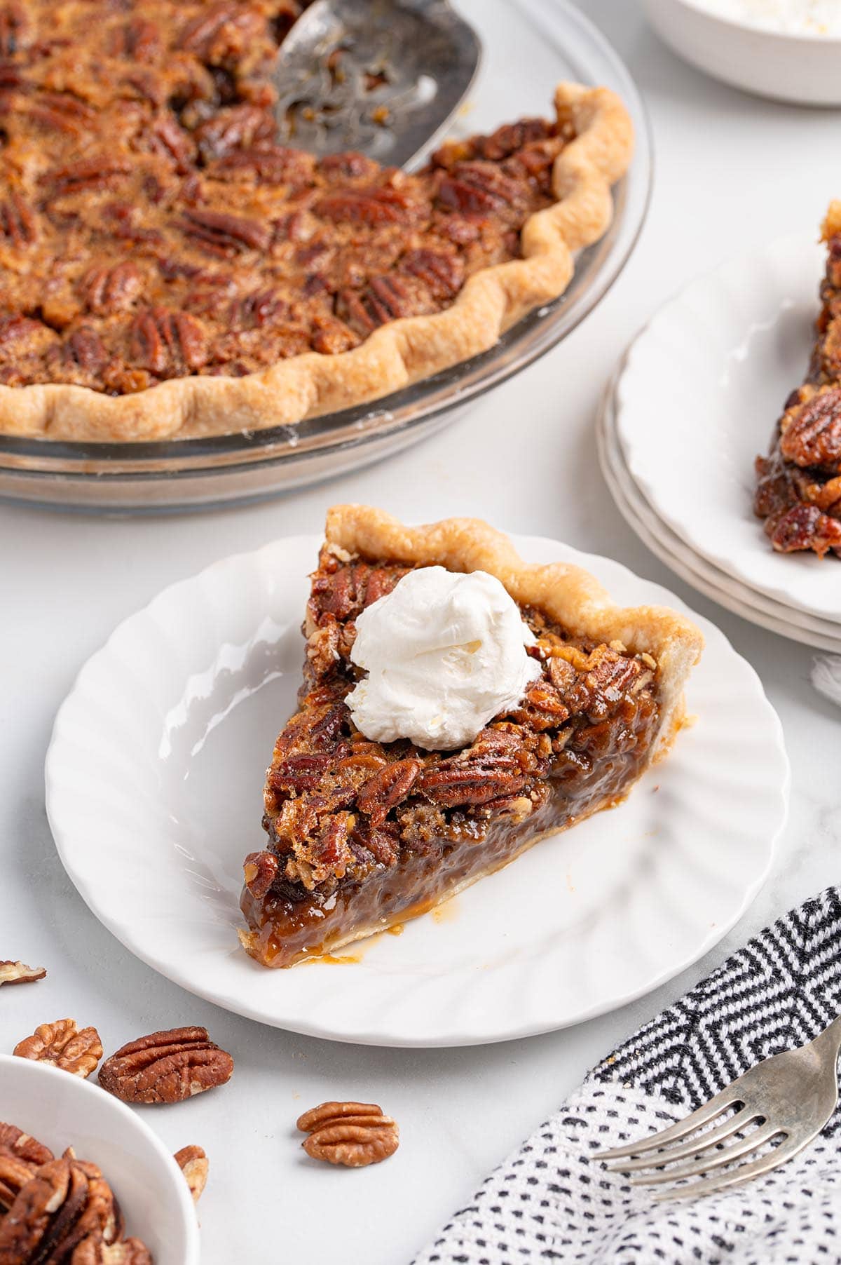 Slice of pecan pie with whipped cream on a plate, whole pie in background.