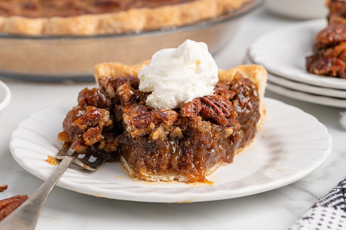 Slice of pecan pie topped with whipped cream on a white plate with a fork.